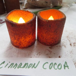 May include: Two small, round, cinnamon and cocoa scented candles with a warm glow. The candles are in a rustic, brown color and have a rough texture.