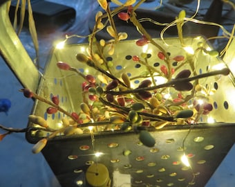 May include: A square metal grater with a perforated surface containing a cluster of artificial berry branches in shades of red, green, and cream, illuminated by thin, flexible copper wire string lights that emit a warm, bright glow throughout.