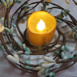 May include: A decorative candle with a faux flame sits within a wreath of brown twigs and white and teal teardrop-shaped beads. The candle has a warm, yellow glow, creating a cozy ambiance. The wreath is placed on a white surface.