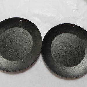 May include: Two black, round metal candle holders with a textured surface. Each has a small hole near the edge. The candle holders are designed to hold candles and add a decorative touch to any space.