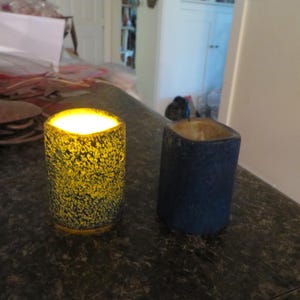May include: Two decorative candle holders. One is lit, with a yellow glow, and has a textured, speckled surface. The other is unlit, blue, and has a similar textured surface. Both are cylindrical with square tops.