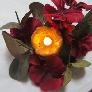 May include: A small, round, orange candle with a white flame sits in the center of a red and green floral arrangement. The flowers are made of fabric and are arranged in a circle around the candle.