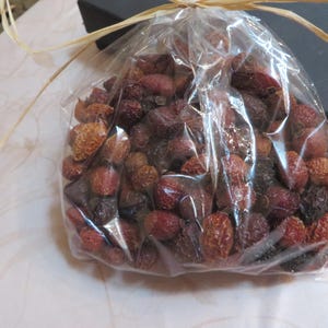 May include: A clear plastic bag filled with dried red and dark brown rose hips. The bag is gathered at the top and tied with a thin, light tan raffia ribbon. The contents consist of numerous small, wrinkled, spherical botanical fruits.