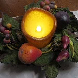 May include: A lit candle in a brown glass jar surrounded by green leaves, berries, and small fruit. The candle is surrounded by a decorative arrangement of fall foliage.