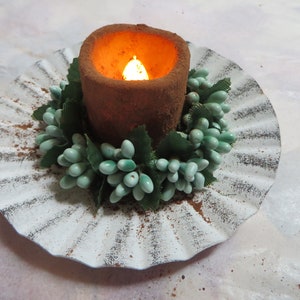 May include: A brown candle with a lit flame sits on a white, scalloped plate. The candle is surrounded by a green garland of artificial berries.