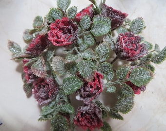 May include: A cluster of artificial dark red roses with green leaves, all covered in a fine layer of white, crystalline frost. The arrangement features multiple blooming flower heads and serrated foliage, creating a dense, textured botanical display.