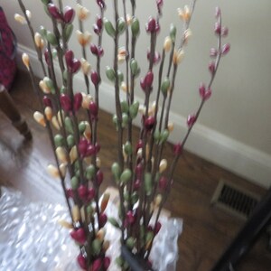 May include: A bundle of artificial floral stems with small, colorful buds. The buds are in shades of burgundy, green, and cream, attached to brown stems. The arrangement is likely for home decor.