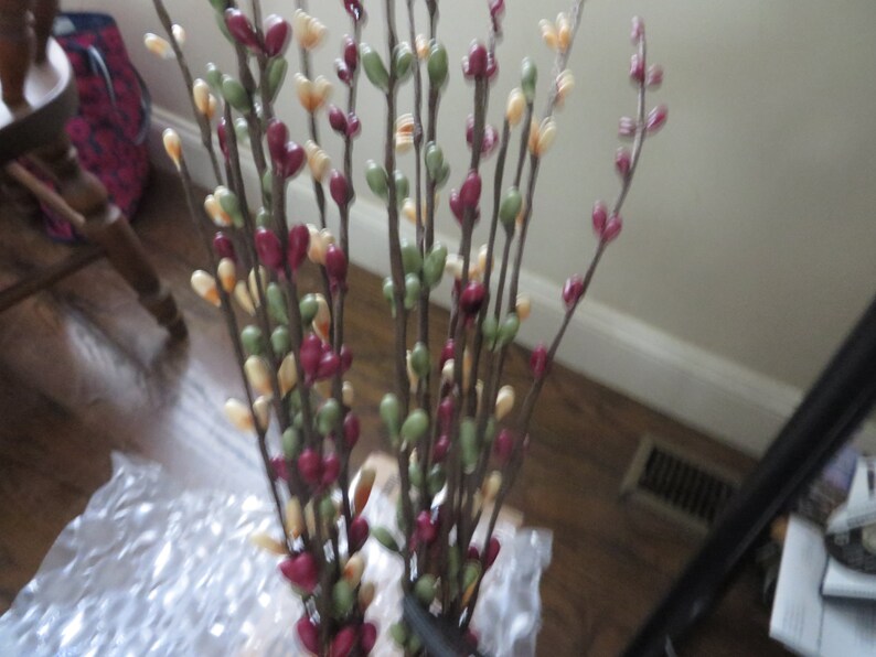 May include: A bundle of artificial floral stems with small, colorful buds. The buds are in shades of burgundy, green, and cream, attached to brown stems. The arrangement is likely for home decor.