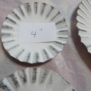 May include: White, ruffled decorative plates with a distressed black edge. One plate has a paper label with the number "4" written on it. The plates are approximately 4 inches in diameter.