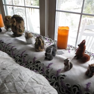 May include: A white tablecloth with a black and purple lace trim, decorated with small stuffed animals, including squirrels, owls, and a fox. Two orange candles are on the table, one in front of a window.