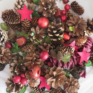 May include: A collection of natural brown pinecones mixed with red decorative stars, shiny red spherical ornaments, red jingle bells, and small red berries. Green leaves are interspersed throughout the arrangement of various sizes.