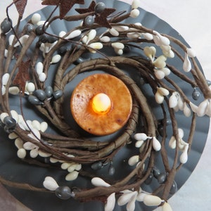 May include: A brown twig wreath with white berries and gray berries surrounding a lit candle in a brown holder.