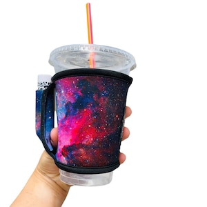 May include: A black and blue galaxy print insulated cup holder with a clear plastic cup and a pink and orange straw.