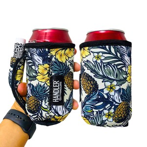May include: Two white and blue tropical print neoprene can coolers with a black handle. The can coolers feature pineapples, flowers, and leaves. The text "HANDLER @LITHANDLERS" is printed on the side of one cooler.