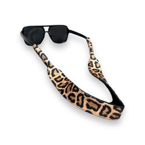 May include: Black sunglasses with a leopard print strap. The sunglasses have a square frame and dark lenses. The strap is tan with black spots. The strap is designed to hold the sunglasses around the neck.