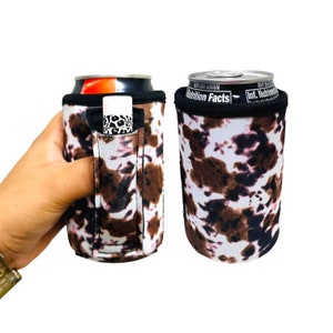 May include: Two can coolers with a cow print design. One cooler has a lip balm in a small pocket. The coolers are white, brown, and black. One can has the words "Nutrition Facts" printed on it.