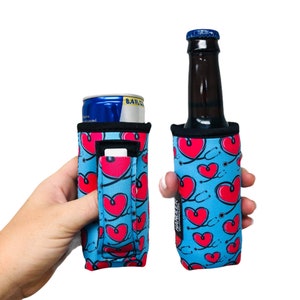 May include: Two blue and pink neoprene can coolers with a stethoscope and heart pattern. The cooler on the left is holding a can of energy drink. The cooler on the right is holding a brown glass bottle. Both coolers have a black strap and a pocket on the side.