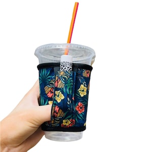 May include: A black neoprene cup holder with a tropical floral print. The holder has a finger loop and a slot for a straw. The cup holder is on a clear plastic cup with a straw.