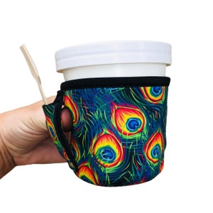 May include: A black and blue neoprene cup holder with a peacock feather design. The holder has a finger loop and a slot for a straw.