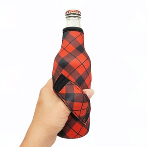 May include: Red and black plaid neoprene bottle cooler with a black strap handle. The cooler is designed to fit a standard-sized bottle.