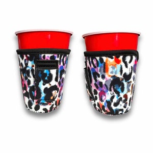 Puede incluir: Dos vasos de plástico rojos se colocan en fundas para vasos con estampado de leopardo colorido. Las fundas tienen un ribete negro y un pequeño bolsillo. El estampado de leopardo presenta manchas azules, moradas, rosas y naranjas sobre un fondo blanco.