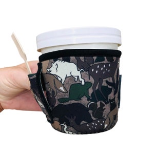 May include: A brown and black insulated cup sleeve with a pattern of various animals, including a deer, a bear, a beaver, and a fox. The sleeve has a pocket on the side for a spoon or straw.