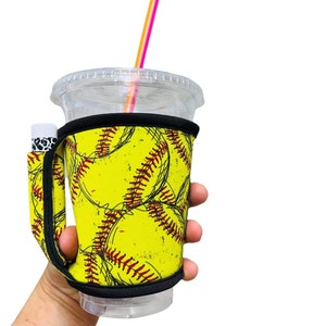 Puede incluir: Un portavasos de neopreno amarillo y negro con un patrón de softball. El portavasos tiene un asa y una correa para llevar. Un vaso de plástico transparente con una pajita rosa y naranja está dentro del portavasos.