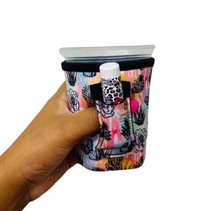 Puede incluir: Una funda de taza colorida con un estampado de cactus y flores. La funda tiene un bolsillo negro que contiene un bálsamo labial y un pompón rosa. La funda está diseñada para sostener una taza con tapa.