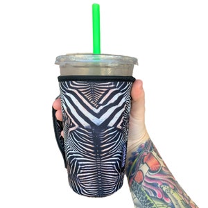 Puede incluir: Un portavasos aislado con estampado de cebra en blanco y negro con una pajita verde que sobresale de un vaso de plástico transparente. El portavasos tiene un asa en el lateral.