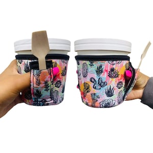 May include: Two white cups with black lids and a cactus print neoprene sleeve. The sleeves have a built-in slot for a spoon or straw. One sleeve has a wooden spoon in the slot.