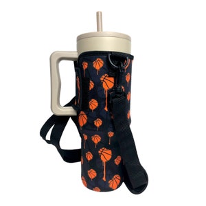 May include: A black and orange insulated tumbler with a basketball print and a black strap. The tumbler has a handle and a straw.