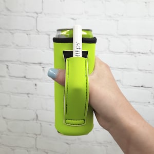 May include: A bright green neoprene can cooler with a black strap and a white marker tucked into the strap. The cooler is being held by a hand.
