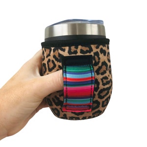 Puede incluir: Un portavasos aislado con estampado de leopardo y un asa de rayas de colores. El portavasos está hecho de neopreno y tiene una cremallera.