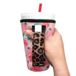 Puede incluir: Un portavasos de neopreno con estampado floral rosa y blanco con un asa de estampado de leopardo. El portavasos tiene una ranura para una pajita y un pequeño bolsillo para un bálsamo labial u otro artículo pequeño.