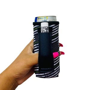 May include: A black and white can cooler with a green and white American flag design. The cooler has a black strap and a pocket for a lip balm. The cooler is holding a can of beer.