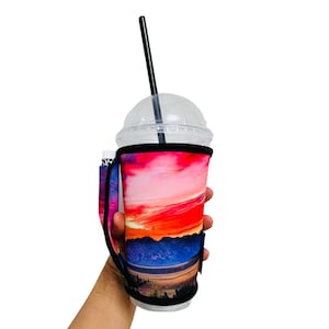 May include: A reusable cup sleeve with a colorful mountain sunset design. The sleeve has a handle and a black strap for carrying.