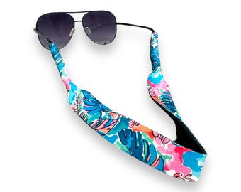 May include: Aviator sunglasses with a colorful strap featuring a tropical floral pattern in shades of blue, pink, and green. The strap is designed to hold the sunglasses around the neck.