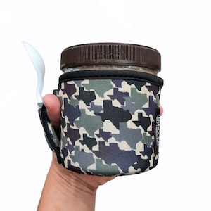 May include: A brown glass jar with a black lid and a camouflage-patterned neoprene sleeve. The sleeve has a white plastic spoon attached to it and features a repeated pattern of the state of Texas.