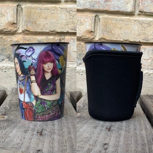 Puede incluir: Un vaso alto y cilíndrico con un gráfico colorido de dos personajes con cabello morado y azul. El vaso tiene un borde negro y una funda de neopreno negra. Los personajes son de la película de Disney, Descendants.