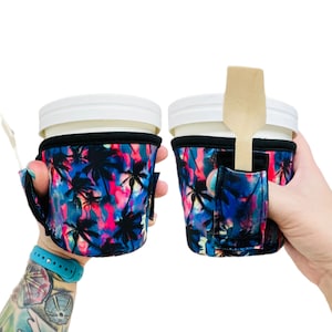 May include: Two insulated cup holders with a tropical palm tree print. The holders are made of neoprene and have a built-in slot for a spoon or straw. The holders are shown on a person's hands.