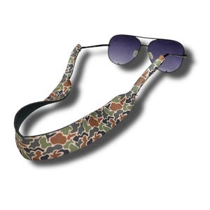 Puede incluir: Una correa para gafas con estampado de camuflaje hecha de material suave y flexible. La correa está diseñada para sujetar las gafas de forma segura y evitar que se caigan.