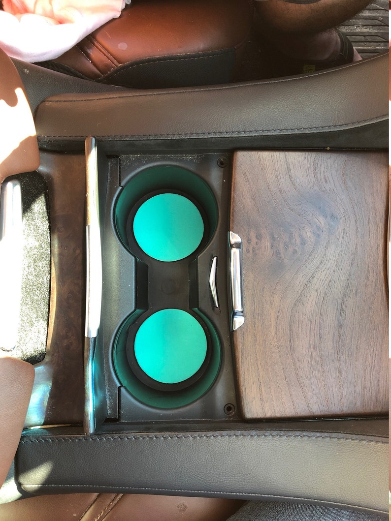 May include: A car's center console with two cup holders, each with a bright teal insert. The console is black with a wood-grain panel and chrome accents. The surrounding upholstery is brown and black leather.