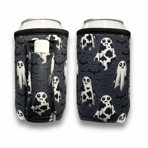 May include: A black and white can cooler with a ghost and bat pattern. The cooler has a pocket for a lip balm or other small item.