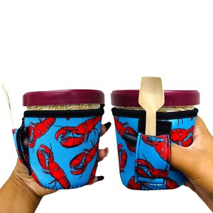 May include: Two blue and red lobster print insulated cup holders with a built-in spoon holder. One cup holder is being held by a hand.