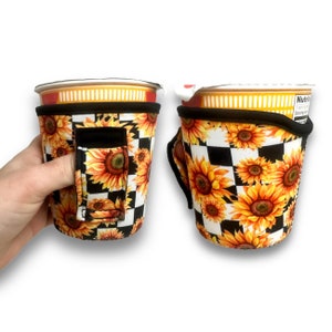 Puede incluir: Un portavasos a cuadros blanco y negro con un patrón de girasoles. El portavasos tiene un asa negra y está diseñado para adaptarse a una taza de tamaño estándar.