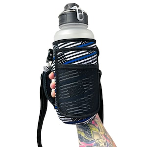 Puede incluir: Un portador de botella de agua negro con un diseño de bandera estadounidense de línea azul delgada. El portador tiene un bolsillo de malla y una correa para el hombro.