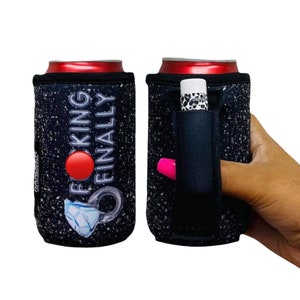 May include: Black and white can cooler with the text "F*cking Finally" and a diamond ring graphic. The cooler has a built-in pocket for a lip balm or other small item.