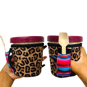 Puede incluir: Dos vasos aislados con tapas, uno con una cuchara de madera unida a una correa de rayas de colores. Ambos vasos están cubiertos con una tela de estampado de leopardo marrón y negro.