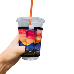 May include: A colorful mountain sunset design printed on a neoprene cup holder with a black strap and a slot for a straw. The holder is on a clear plastic cup with a straw.