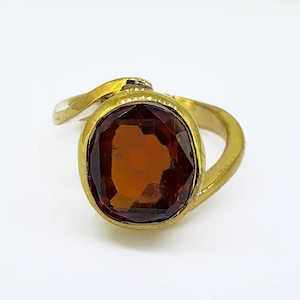 May include: A gold-toned ring featuring a large, oval-shaped, faceted amber-coloured gemstone set in a smooth metal bezel. The band has a curved, asymmetrical design that wraps around the central stone, creating a continuous, polished metallic frame.